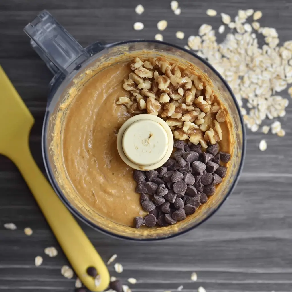 Smooth chickpea blondie batter in a food processor after blending, with dark chocolate chips and walnuts being folded in with a spatula.